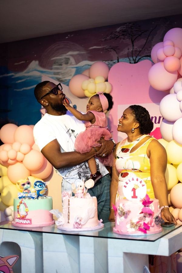 A dad holding his baby daughter beside three pink first-birthday cakes